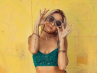 stylish woman wearing green sports bra drawing attention to her green glasses. Posing in front of a yellow wall.